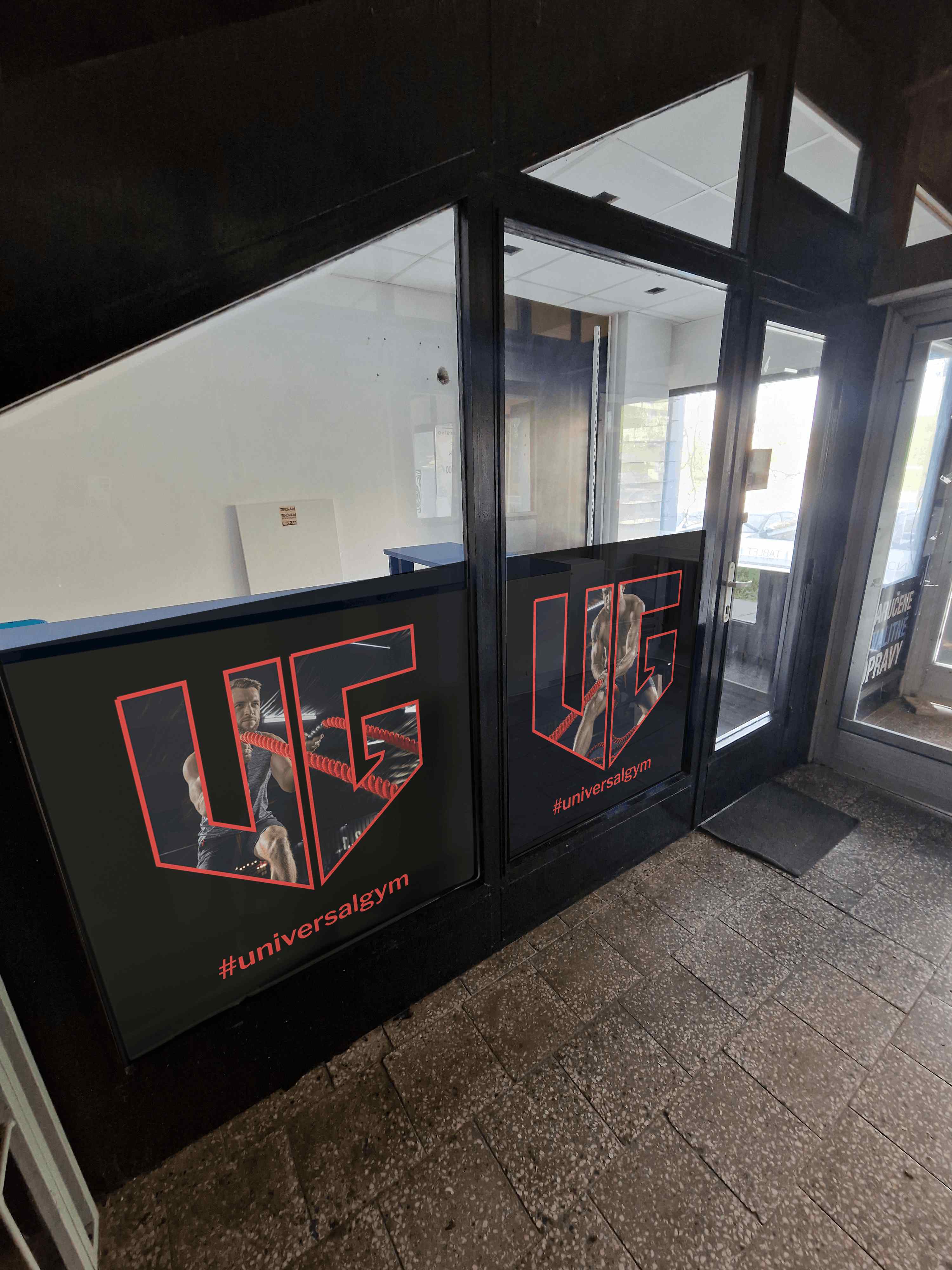 Čierny polep sklenených plôch pri vstupe do Universal Gym s červeným logom UG, fotografiami športovcov a názvom fitness centra.