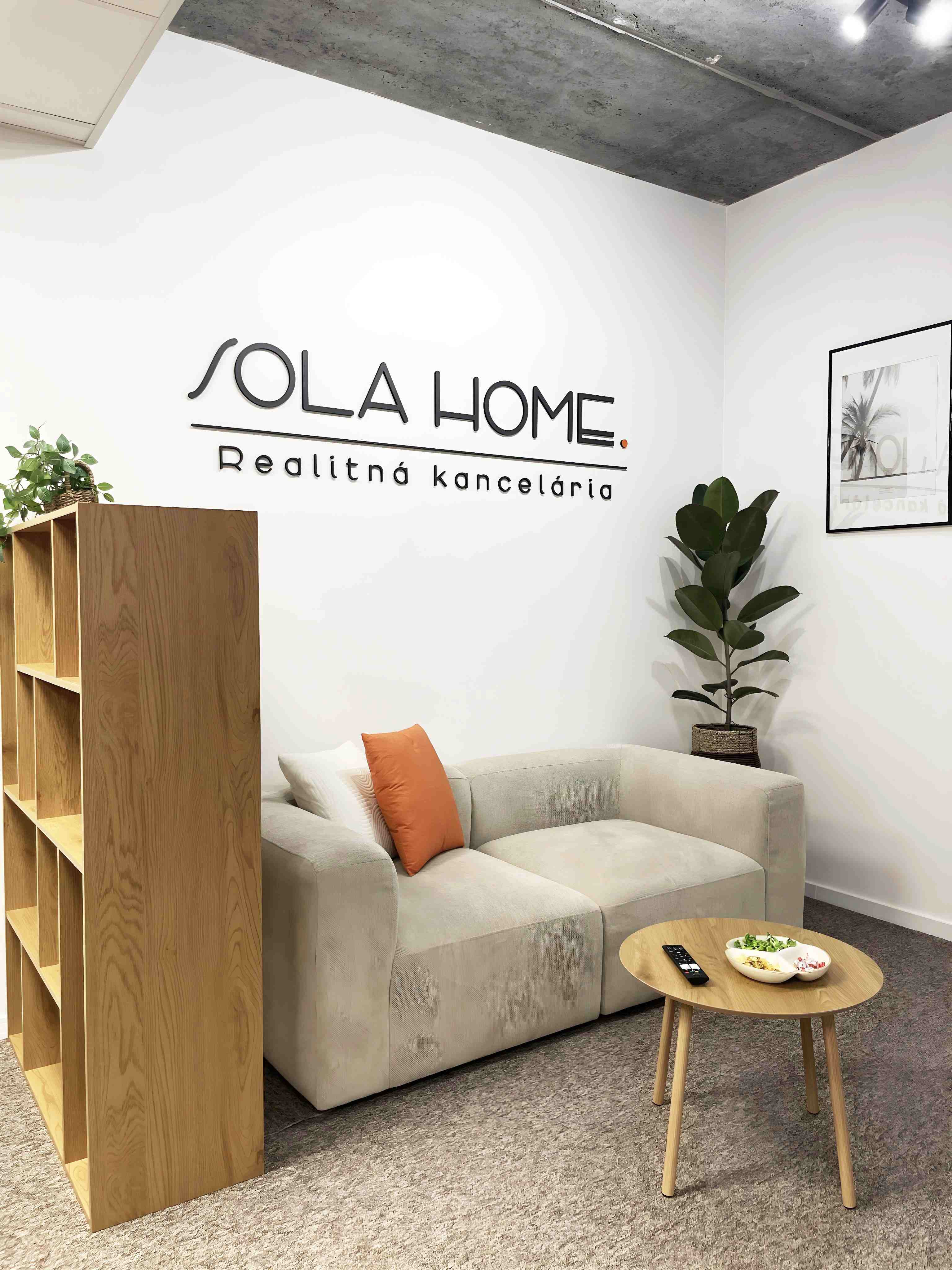 Priestorové logo SOLA HOME na bielej stene nad pohovkou v realitnej kancelárii.