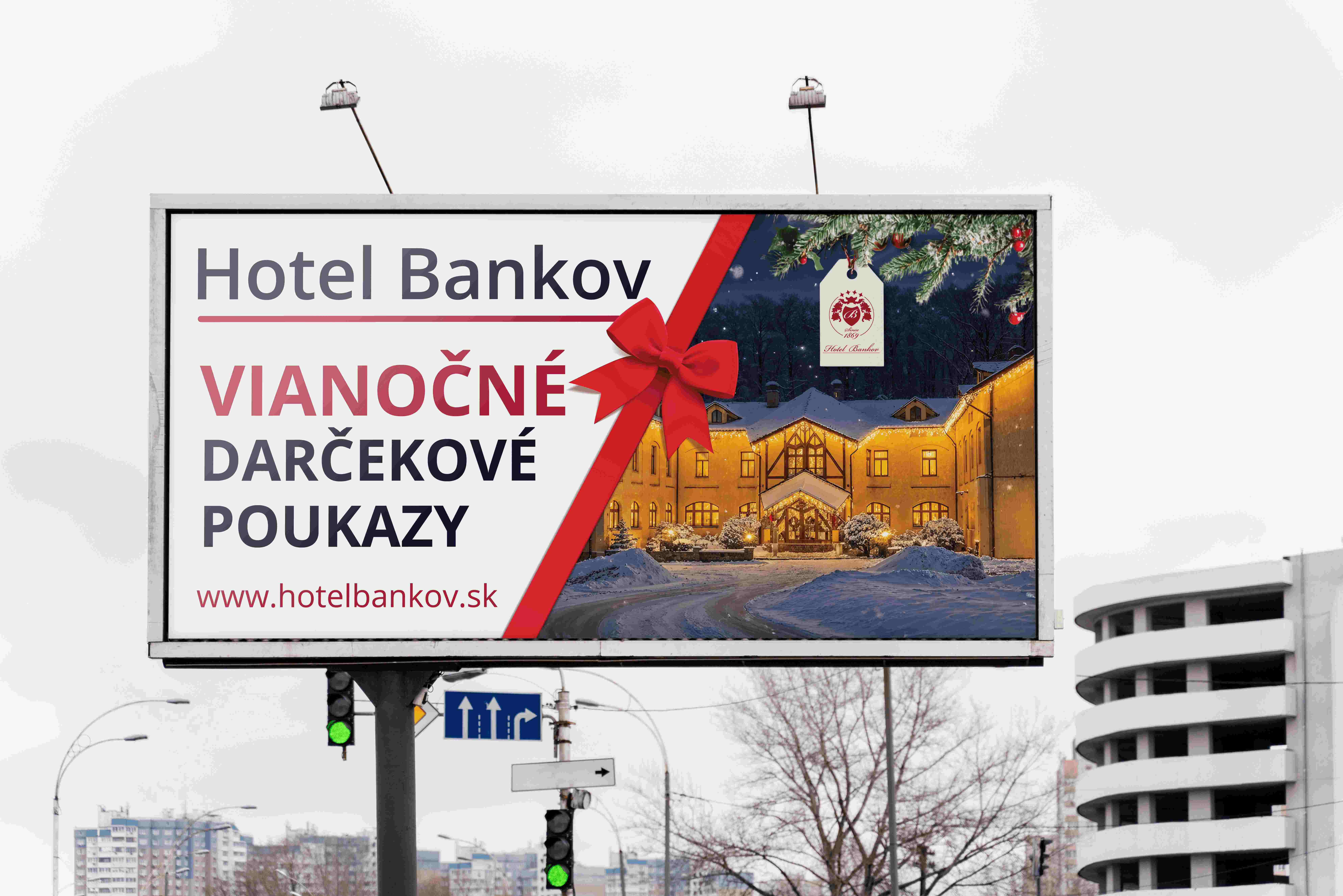 Vianočný reklamný billboard Hotela Bankov s fotografiou zasneženého hotela, červenou mašľou a ponukou darčekových poukazov.