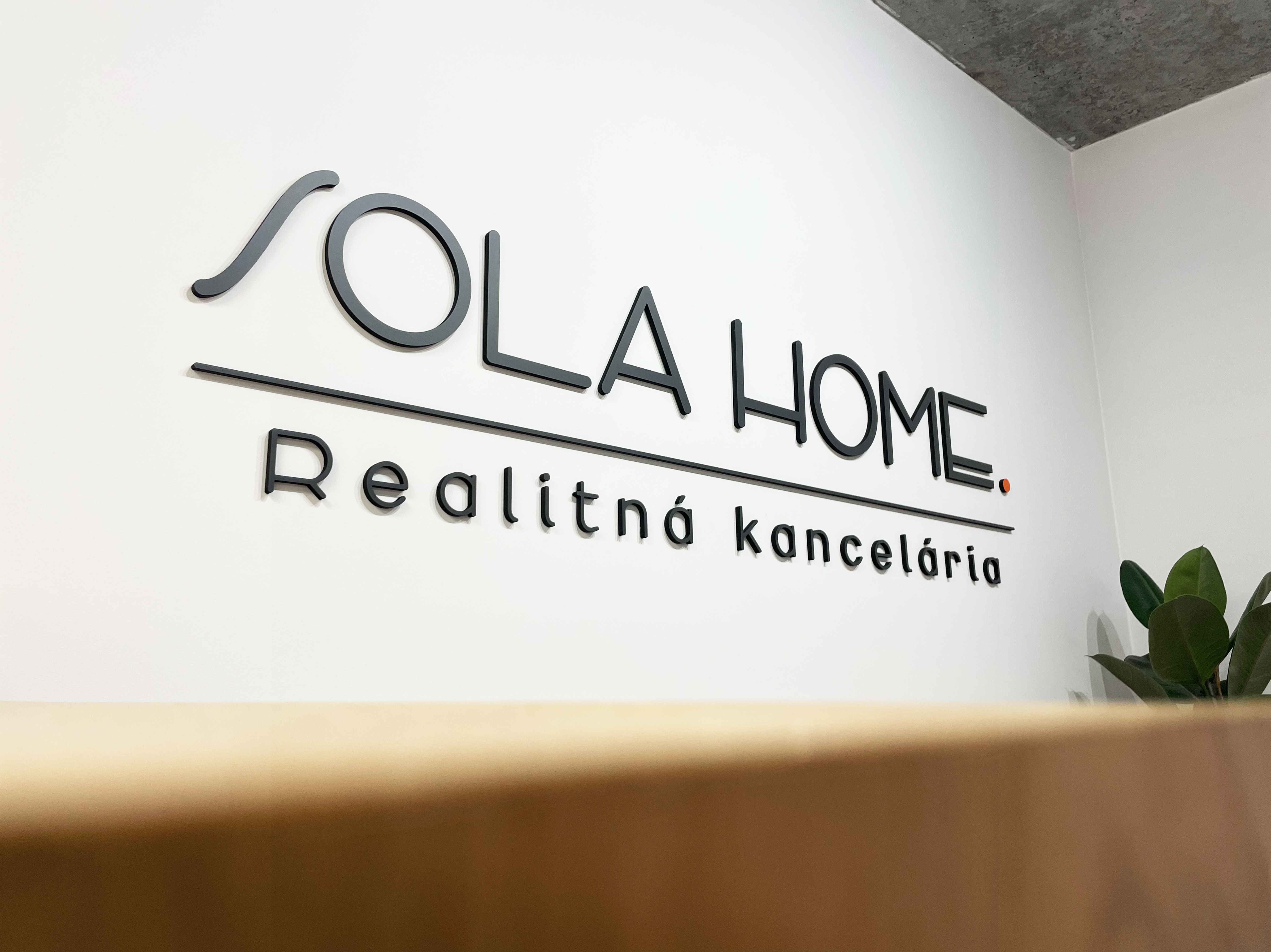Detail sivého loga SOLA HOME s oranžovou bodkou a nápisom Realitná kancelária na stene.