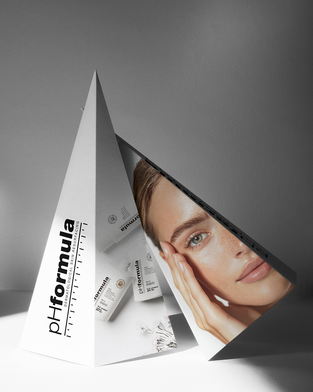 Biely trojuholníkový reklamný stojan pHformula s čiernym logom, fotografiou ženskej tváre a kozmetickými produktmi.