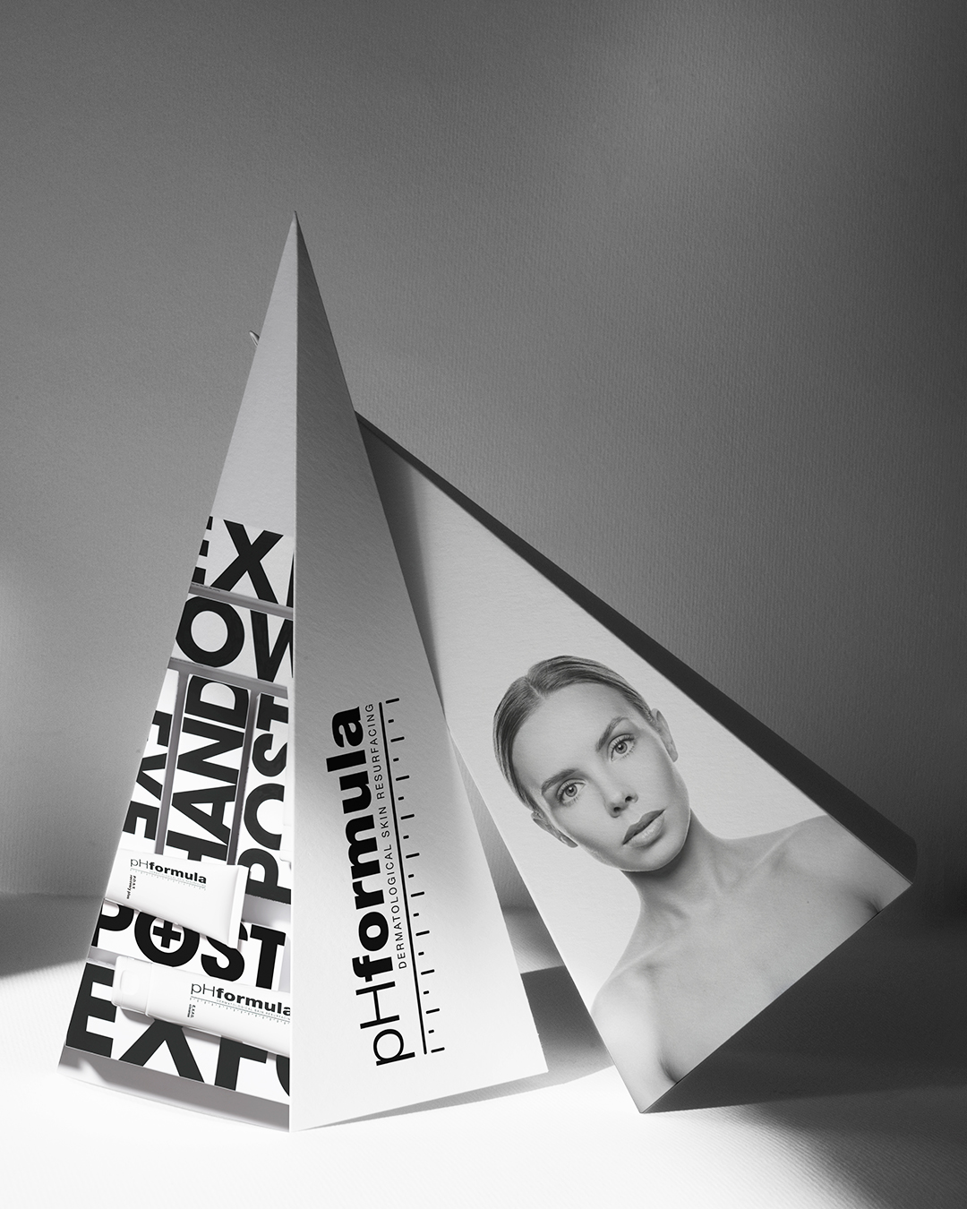 Biely trojuholníkový prezentačný stojan pHformula s čiernym logom, výraznou typografiou, fotografiou modelky a kozmetickými produktmi.
